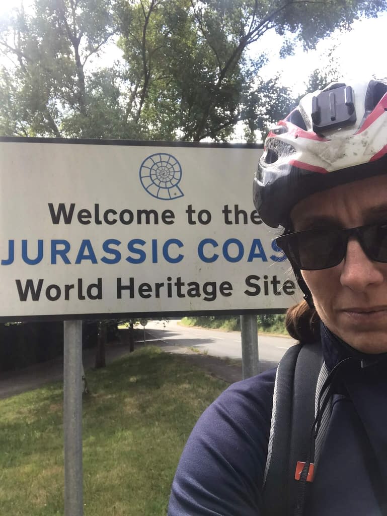 Woman in bike helmet standing in front of a sign that says Welcome to the Jurassic Coast World Heritage Site 