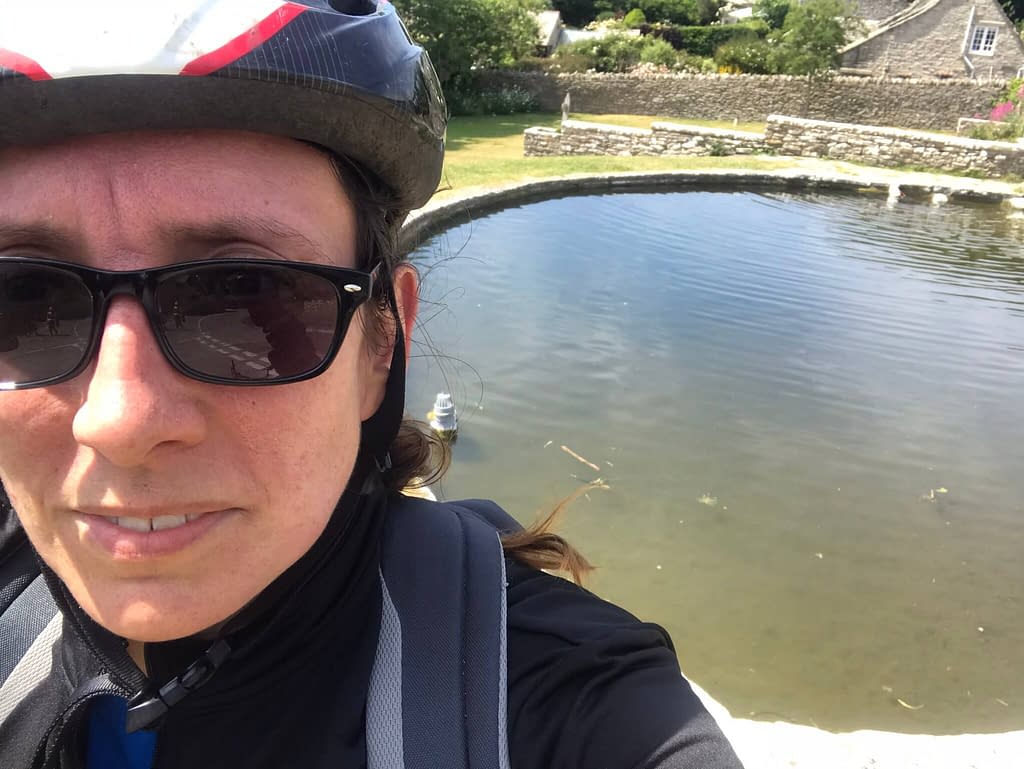 Woman in bike helmet smiling at camera wearing sunglasses in front of a duck pond in Worth Matravers 