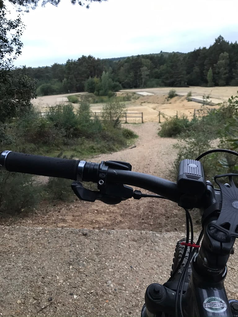 Mountain bike handlebars overlooking view of sandy ground with jumps and tress in background 