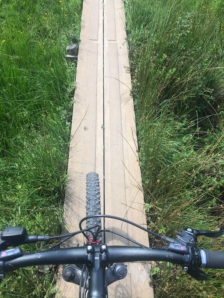 Two wooden boards making a path over long grass being ridden on a mountain bike with wheel and handlebars 