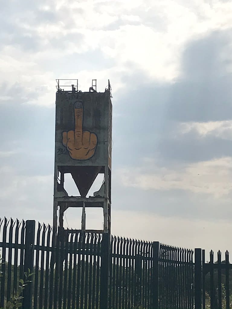 Water tower above metal fencing in industrial estate depicting graffiti of middle finger raised