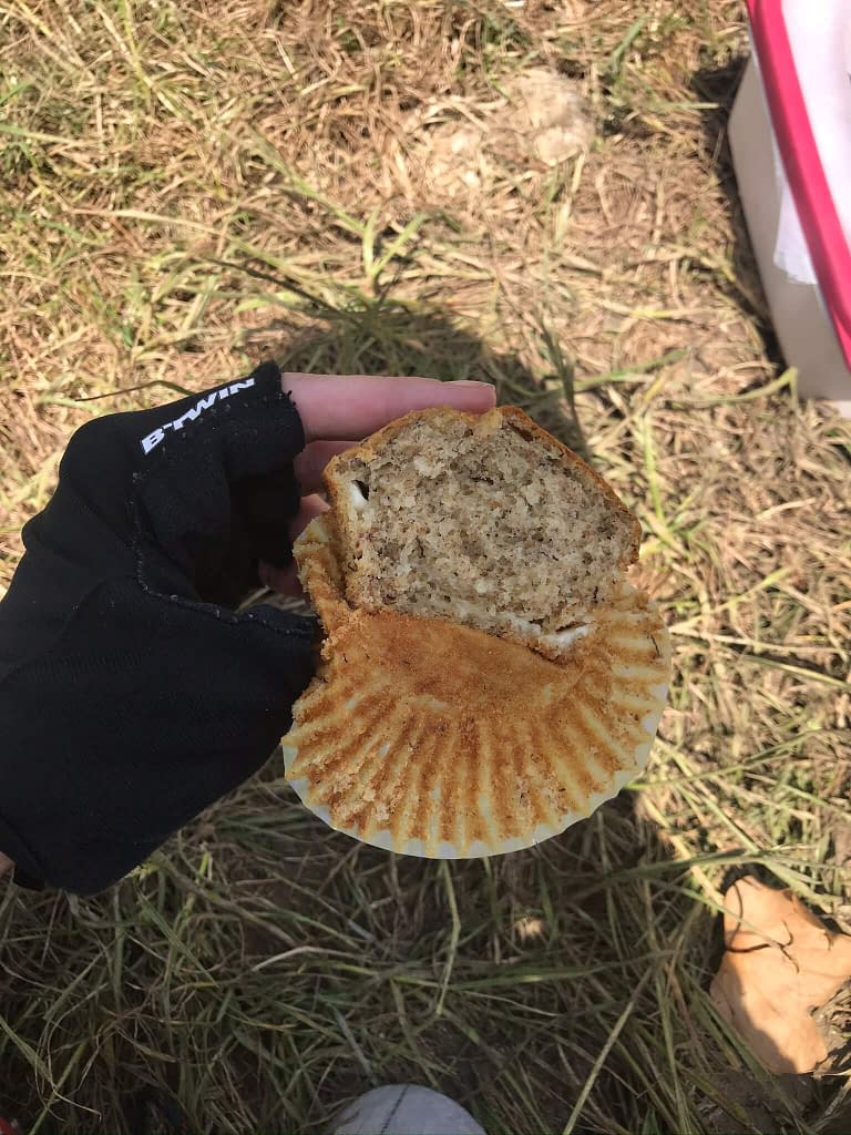 Hand in cycling glove holding a half eaten banana muffin sitting on a grassy field 